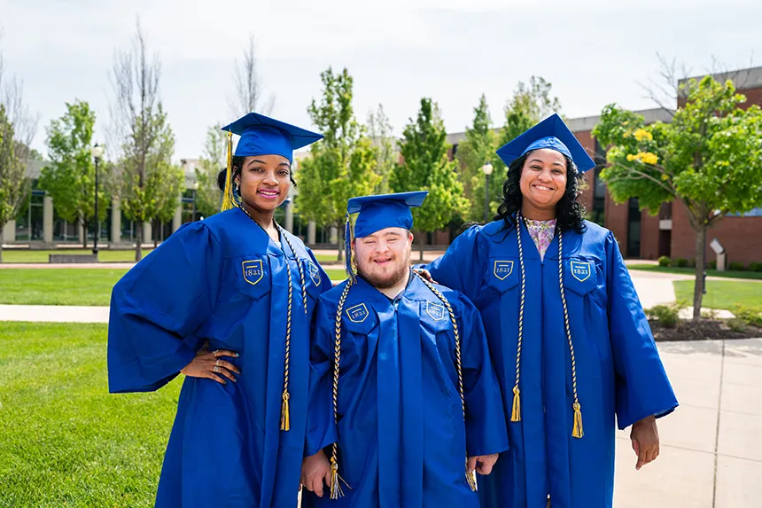 Three grads in blue caps and gowns from the integrated professional studies program pose for a photo on campus