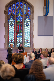 Vincent Starkey speaking at a podium in front of a stained glass window