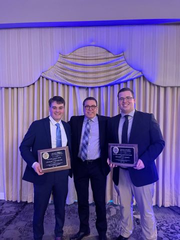 Three people pose for a photo, two of them hold awards. 