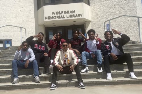 Seven members of Kappa Alpha Psi Fraternity, wearing their fraternity shirts, sit on the steps of the library displaying the fraternity's hand sign