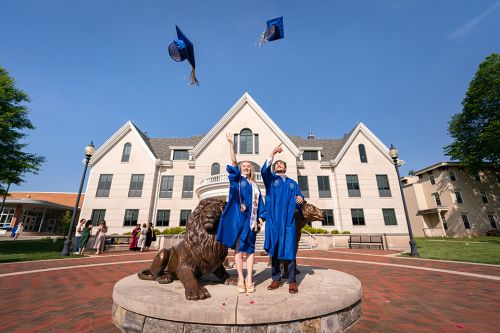 Two graduates in blue gowns tossing their grad caps in the air at the Pride lion statues
