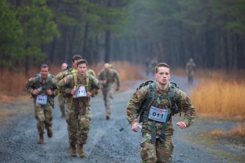 Group of people in Army fatigues and backpacks running along a road in a wooded area
