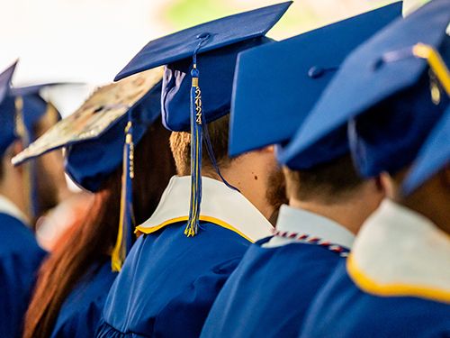 Back of a row of graduates in blue caps and gowns, with the center grad’s tassel of 2024 visible