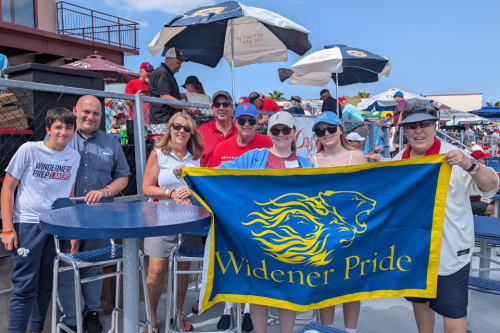 A group of alumni pose with a blue Widener Pride flag at the ballpark