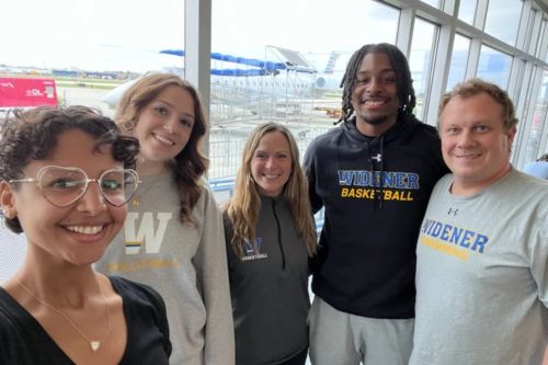 A group of student athletes and athletic staff pose for a photo at the airport before their conference trip.