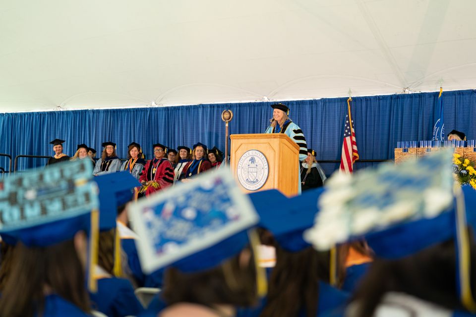President Robertson, in full academic regalia, stands at the podium speaking; in the foreground are blurred blue caps